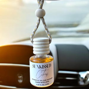 May include: A white and gold car air freshener with a clear glass bottle filled with a yellow liquid. The label reads "Sunkissed Citrus Aromatic Oil Car Diffuser Made with Love 0.27 fl oz | 8 ml".