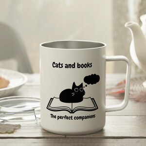 Könnte beinhalten: Weiße Reise-Thermoskanne aus Edelstahl mit einer schwarzen Katze, die auf einem Buch sitzt, und einer Gedankenblase über der Katze. Der Text auf der Tasse lautet "Cats and books The perfect companions".