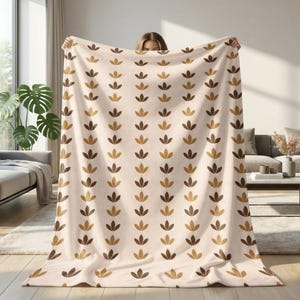 May include: A cream-coloured blanket featuring a repeating pattern of stylised brown and gold leaf shapes. The blanket is held up, showcasing its soft texture and design. A cosy home decor item.
