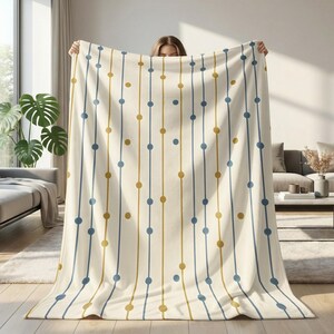 May include: Cream-colored blanket with a repeating pattern of vertical lines. Each line alternates between blue and gold, with evenly spaced dots of the same color. The blanket is held up, showcasing its design.