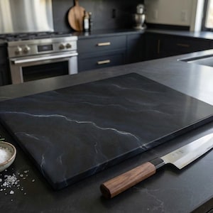 May include: A black rectangular chopping board with white veining is on a countertop next to a knife with a wooden handle and a small bowl of salt. The kitchen setting includes a stainless steel stove and sink.