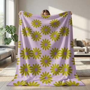 Puede incluir: Una manta lila con un patrón repetido de grandes flores de margarita amarillas y moradas. La manta se sostiene, mostrando el diseño. La manta es un artículo de decoración del hogar suave y acogedor.