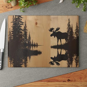 Peut inclure: Une planche à découper en bois avec une silhouette d'un élan et des pins reflétés dans un lac. La planche a une finition en bois naturel.