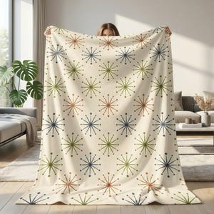 May include: Cream-coloured blanket featuring a repeating pattern of starburst designs in brown, green, and navy blue. The blanket is held up, displaying its full design and texture. A cosy home decor item.