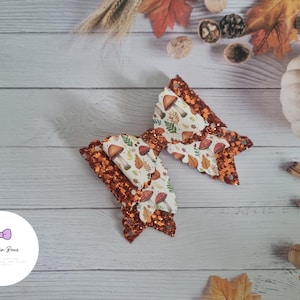 Könnte beinhalten: Eine große Haarschleife mit Herbstthema. Die Schleife hat ein mehrschichtiges Design mit einer glitzernden orangefarbenen Basis und einer gemusterten oberen Schicht mit Pilzen und Blättern. Die Schleife ist von Herbstlaub und einem Kürbis umgeben.