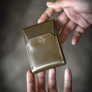 Op de afbeelding: Olijfgroene leren portemonnee met een gestikte rand en een cirkelvormig logo. De portemonnee wordt tussen twee handen gehouden, wat het ontwerp en het vakmanschap laat zien. De portemonnee lijkt nieuw en ongebruikt.