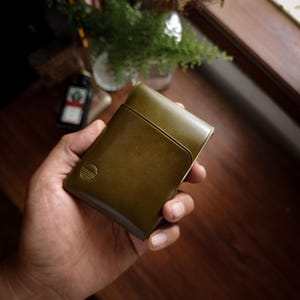 Puede incluir: Cartera de cuero verde oliva con un diseño minimalista. La cartera tiene un pequeño logotipo en relieve y una ranura para tarjetas. La cartera se sostiene en una mano, con una superficie de madera oscura en el fondo.