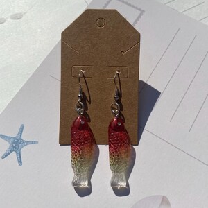 May include: A pair of dangle earrings featuring red, orange, and yellow fish-shaped charms. The fish are translucent and have a textured surface.