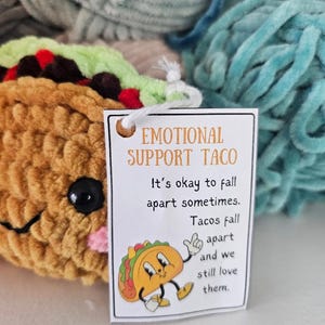 May include: A white card with the text "Emotional Support Taco" and the message "It's okay to fall apart sometimes. Tacos fall apart and we still love them."  A cartoon taco with a happy face is illustrated on the card.