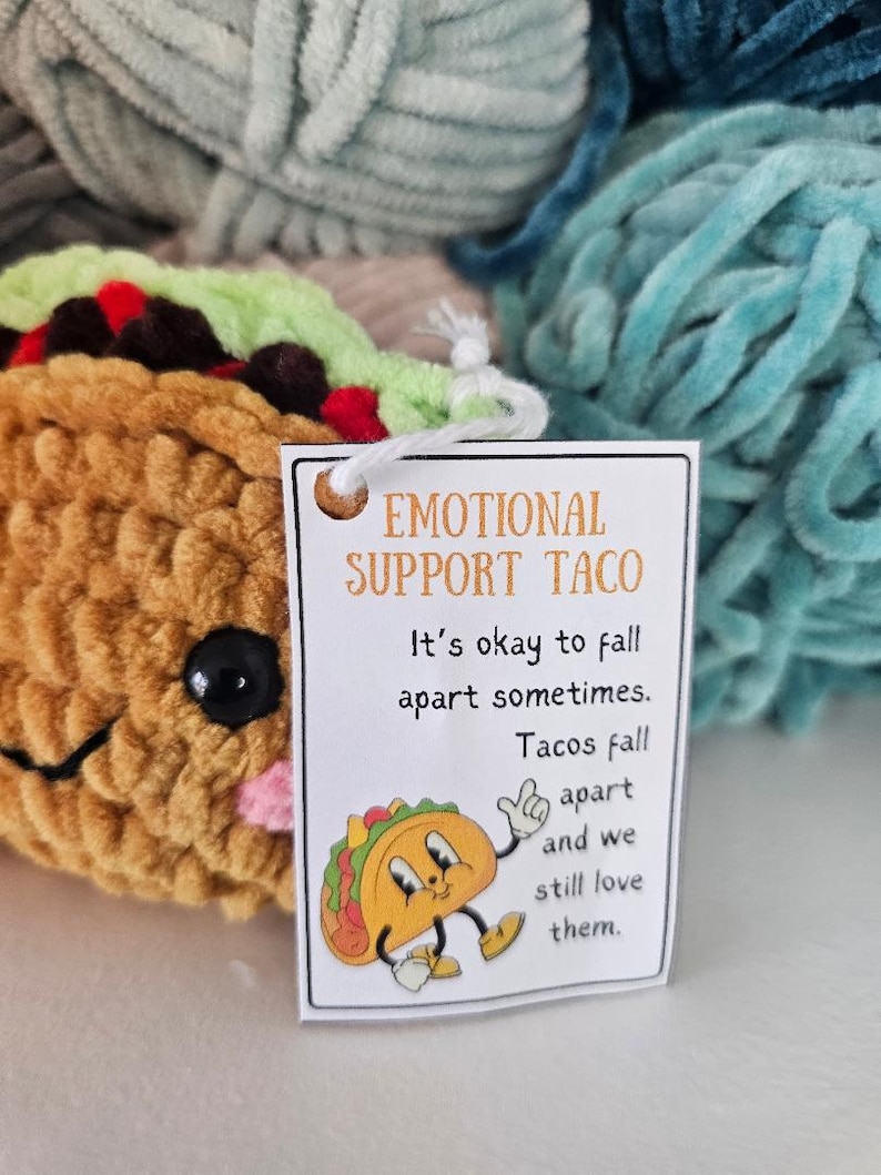 May include: A white card with the text "Emotional Support Taco" and the message "It's okay to fall apart sometimes. Tacos fall apart and we still love them."  A cartoon taco with a happy face is illustrated on the card.