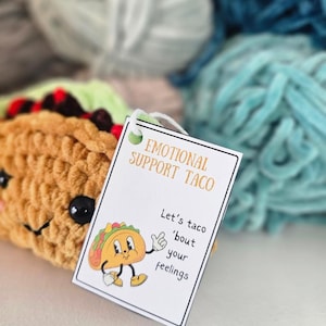 May include: A white tag with the text "Emotional Support Taco" and "Let's taco 'bout your feelings" with a cartoon taco illustration. The tag is placed on top of a crocheted taco plush toy.