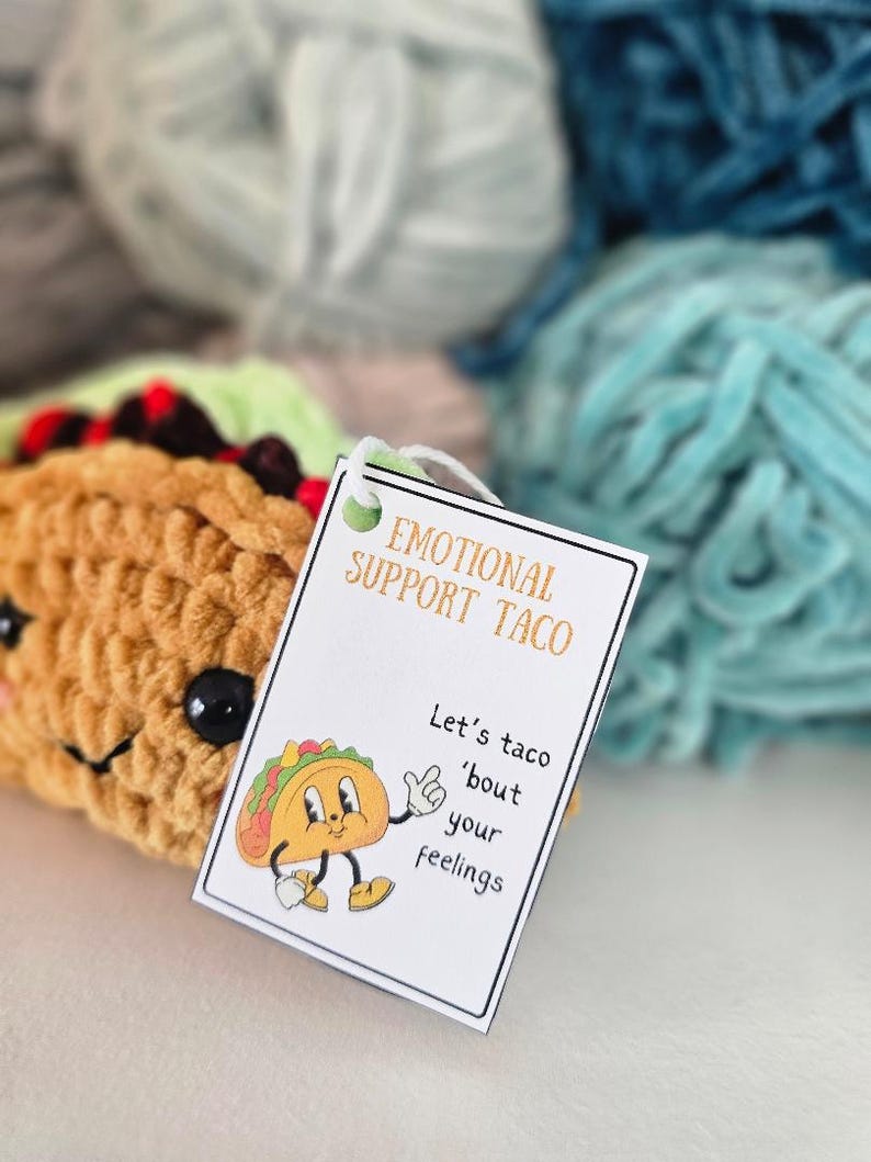 May include: A white tag with the text "Emotional Support Taco" and "Let's taco 'bout your feelings" with a cartoon taco illustration. The tag is placed on top of a crocheted taco plush toy.