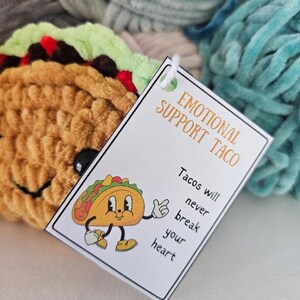 May include: A white card with the text "Emotional Support Taco" and "Tacos will never break your heart". A cartoon taco with a smiling face is on the card. The card is on top of a pile of colorful yarn.