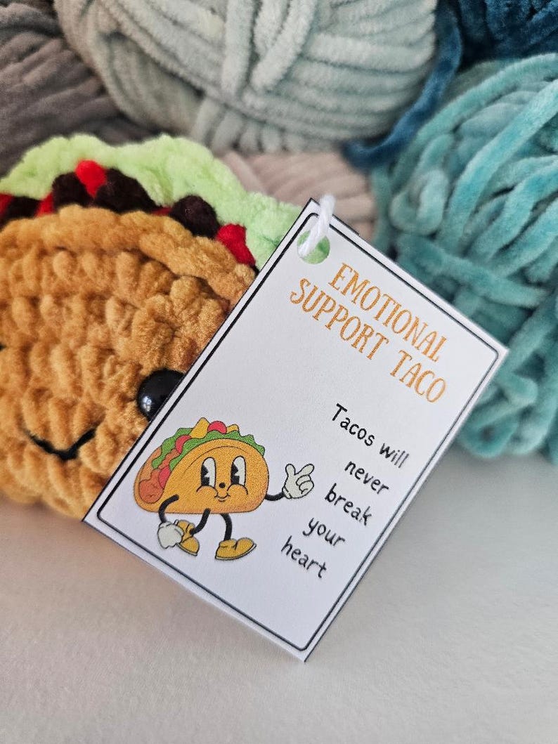 May include: A white card with the text "Emotional Support Taco" and "Tacos will never break your heart". A cartoon taco with a smiling face is on the card. The card is on top of a pile of colorful yarn.
