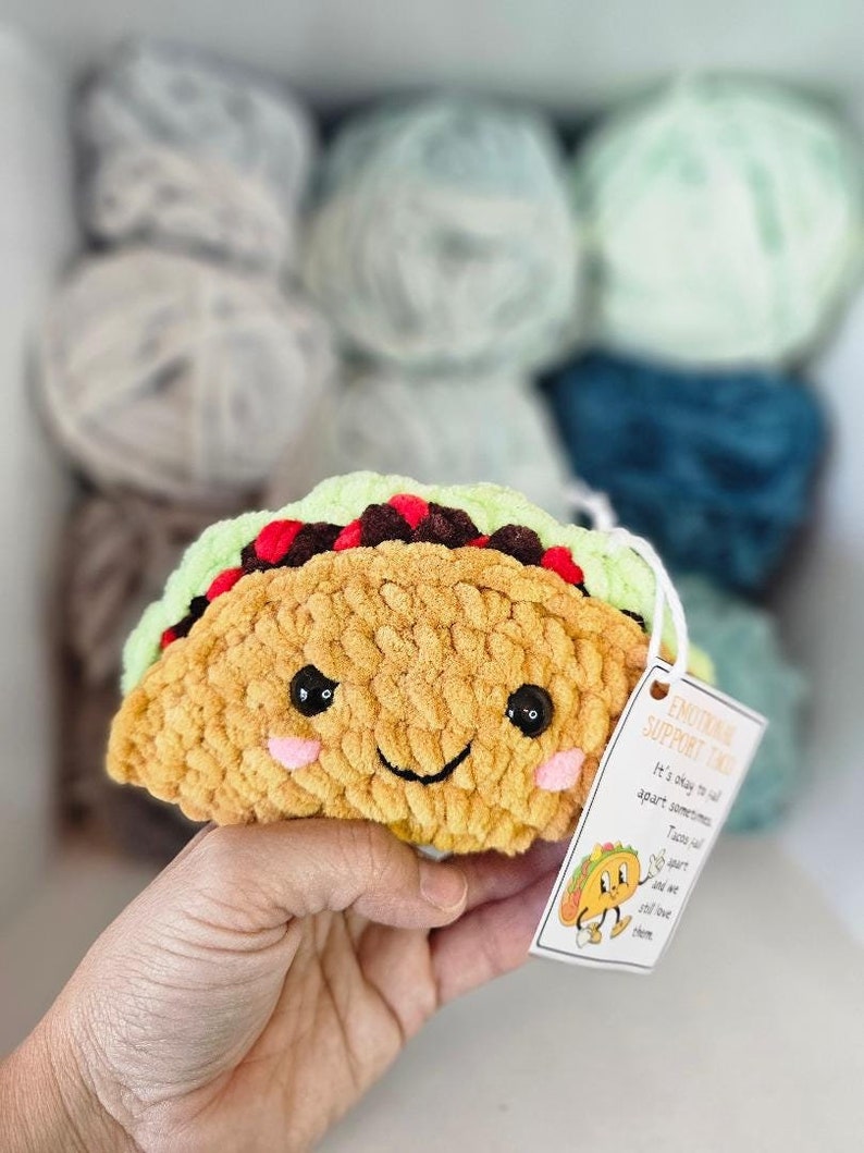 May include: A crocheted taco plush toy with a smiling face and a tag that reads "Emotional Support Taco. It's okay to fall apart sometimes. Taco 'bout good times and we still love them."