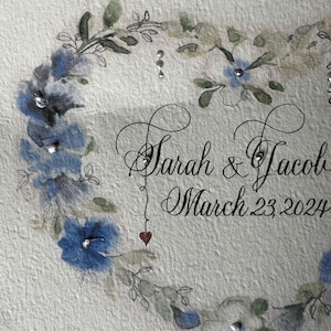 Puede incluir: Una pintura de acuarela de una corona floral con flores azules y hojas verdes. El texto "Sarah & Jacob March 23, 2024" est&aacute; escrito en cursiva.