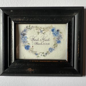 Puede incluir: Una pintura de acuarela enmarcada de un coraz&oacute;n hecho de flores azules con el texto "Sarah & Jacob March 23, 2024" en caligraf&iacute;a negra.