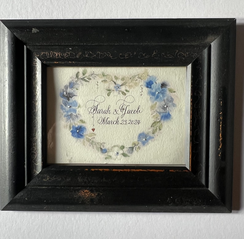 Puede incluir: Una pintura de acuarela enmarcada de un coraz&oacute;n hecho de flores azules con el texto "Sarah & Jacob March 23, 2024" en caligraf&iacute;a negra.