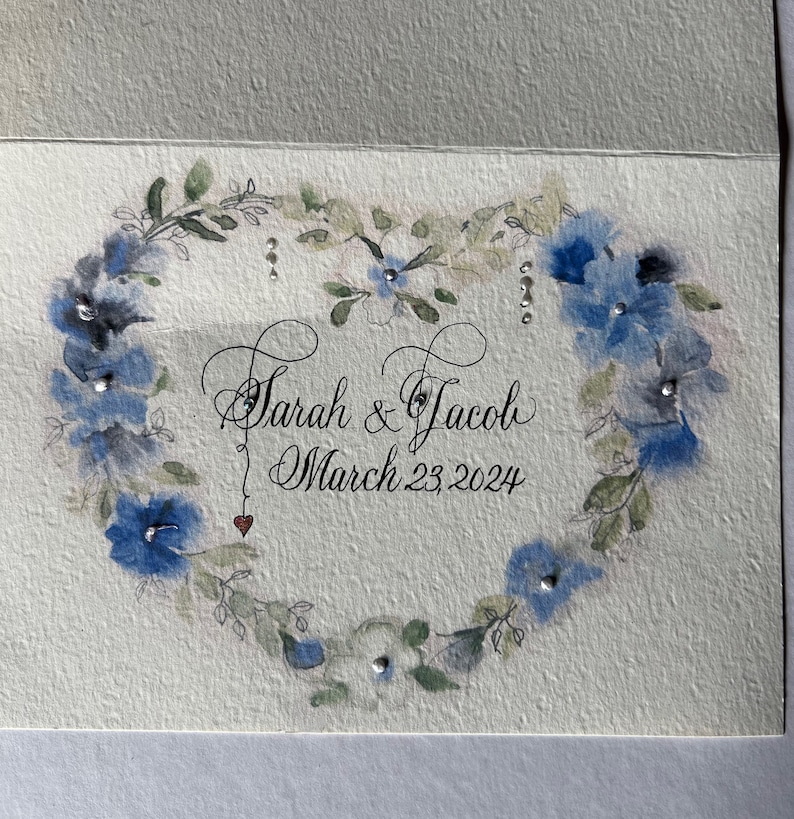Puede incluir: Una ilustraci&oacute;n de acuarela de una corona en forma de coraz&oacute;n de flores azules con acentos plateados. El texto "Sarah & Jacob" y "March 23, 2024" est&aacute; escrito en letra cursiva.