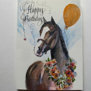 May include: A watercolor painting of a brown and white horse with a brown halter. The horse is wearing a floral garland and has a brown balloon tied to its mane. The text "Happy Birthday" is written in cursive at the top of the card.