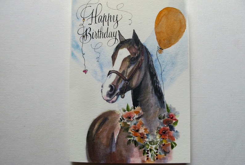 May include: A watercolor painting of a brown and white horse with a brown halter. The horse is wearing a floral garland and has a brown balloon tied to its mane. The text "Happy Birthday" is written in cursive at the top of the card.