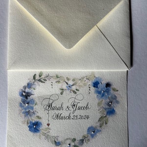 Puede incluir: Una tarjeta blanca con una corona floral azul en forma de coraz&oacute;n. El texto "Sarah & Jacob March 23, 2024" est&aacute; escrito en cursiva negra dentro de la corona.