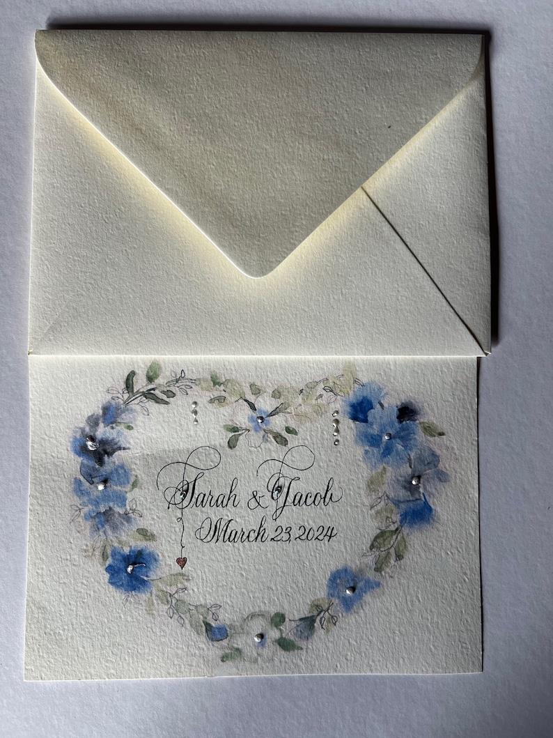 Puede incluir: Una tarjeta blanca con una corona floral azul en forma de coraz&oacute;n. El texto "Sarah & Jacob March 23, 2024" est&aacute; escrito en cursiva negra dentro de la corona.