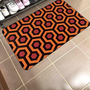 Puede incluir: Felpudo naranja y negro con diseño geométrico. El patrón se asemeja a la alfombra de la película "El Resplandor".