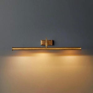 May include: A brass-toned picture light fixture mounted on a dark blue wall. The light has a rectangular backplate and a horizontal bar with a downward-facing light. The light illuminates the wall below.
