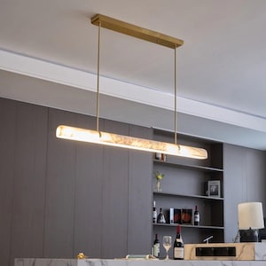 Może przedstawiać: Liniowa lampa wisząca z długim, cylindrycznym, półprzezroczystym białym kloszem o marmurkowej fakturze. Lampa jest zawieszona na prostokątnym mocowaniu sufitowym w kolorze mosiądzu za pomocą dwóch cienkich, pionowych metalowych prętów.