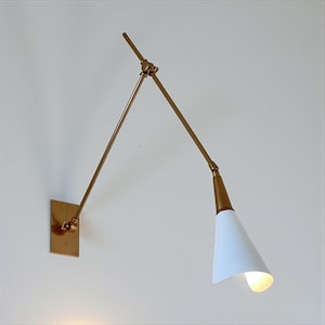 May include: A wall-mounted, adjustable lamp with a gold-toned metal frame and a white conical shade. The lamp features a rectangular base and a pivoting arm, allowing for directional lighting. The shade is angled downward, emitting a warm glow.