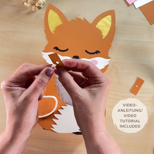K&ouml;nnte beinhalten: Ein Bastelprojekt mit einem Fuchs aus Papier in Orange, Wei&szlig; und Gelb. Der Fuchs hat geschlossene Augen. Der Text "VIDEO ANLEITUNG/ VIDEO TUTORIAL INCLUDED" ist sichtbar. Das Projekt liegt auf einer Holzoberfl&auml;che.
