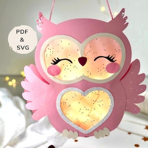 Könnte beinhalten: Eine rosa Papier-Eulenlaterne mit Glitzerakzenten. Die Eule hat große Augen, einen herzförmigen Bauch und eine Schnur zum Aufhängen. Der Text "PDF & SVG" befindet sich in einem weißen Kreis. Die Eule ist von innen beleuchtet.