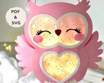 Könnte beinhalten: Eine rosa Papier-Eulenlaterne mit Glitzerakzenten. Die Eule hat große Augen, einen herzförmigen Bauch und eine Schnur zum Aufhängen. Der Text "PDF & SVG" befindet sich in einem weißen Kreis. Die Eule ist von innen beleuchtet.