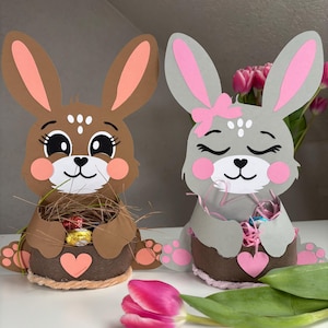 Könnte beinhalten: Zwei Osterhasen-Papierkörbe. Einer ist braun mit Pfirsichakzenten, der andere grau mit rosa Akzenten. Beide haben Körbe mit Ostersüßigkeiten und rosa Herzen gefüllt. Die Hasen haben große Ohren und süße Gesichter.