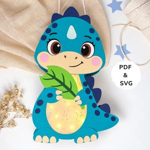 May include: A blue dinosaur paper craft decoration featuring dark blue spots, a light yellow belly with integrated warm LED lights, and a green leaf held in its hands. The design includes a light blue horn, pink cheeks, and the text PDF & SVG.