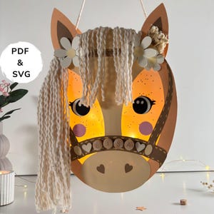 May include: A light brown horse-shaped paper lantern featuring a white yarn mane, white flower accents, and a bridle with the name LISA spelled out on small circular wooden beads. The lantern is illuminated from within and includes PDF & SVG text.