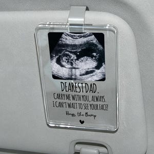 Puede incluir: Un clip rectangular de acrílico transparente para visera de coche con una imagen de ecografía en blanco y negro. El texto dice: DEAREST DAD. CARRY ME WITH YOU, ALWAYS. I CAN'T WAIT TO SEE YOUR FACE! Hugs, the Bump, con un pequeño corazón negro.