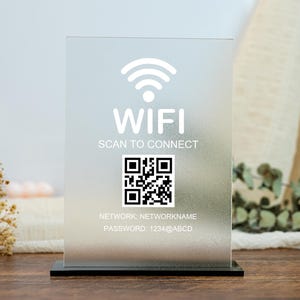 Puede incluir: Un letrero de acrílico transparente con una base negra muestra información de Wi-Fi. El letrero presenta un símbolo de Wi-Fi blanco, la palabra "WIFI" y un código QR. Debajo del código QR se encuentra el nombre de la red y la contraseña.