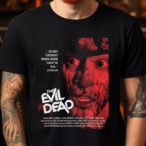 Puede incluir: Una camiseta negra con un gráfico rojo y blanco del póster de la película "The Evil Dead". El póster presenta un primer plano de la cara de un personaje cubierta de sangre y efectos gore.