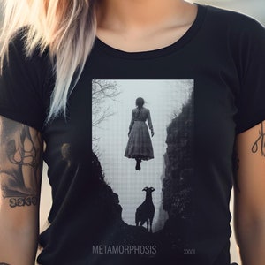 May include: A woman wearing a black t-shirt with a graphic print of a woman in a dress floating above a goat. The text "METAMORPHOSIS" is printed below the graphic. The woman is wearing sunglasses and has blonde hair.