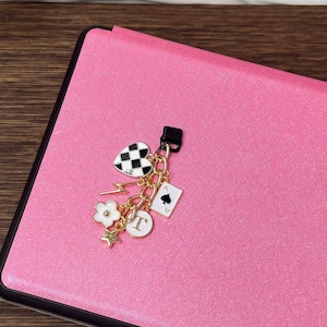 May include: A pink leather notebook with a gold charm attached. The charm features a black and white checkered heart, a gold lightning bolt, a white flower, a gold star, a gold letter 'T', and a playing card with a spade symbol.