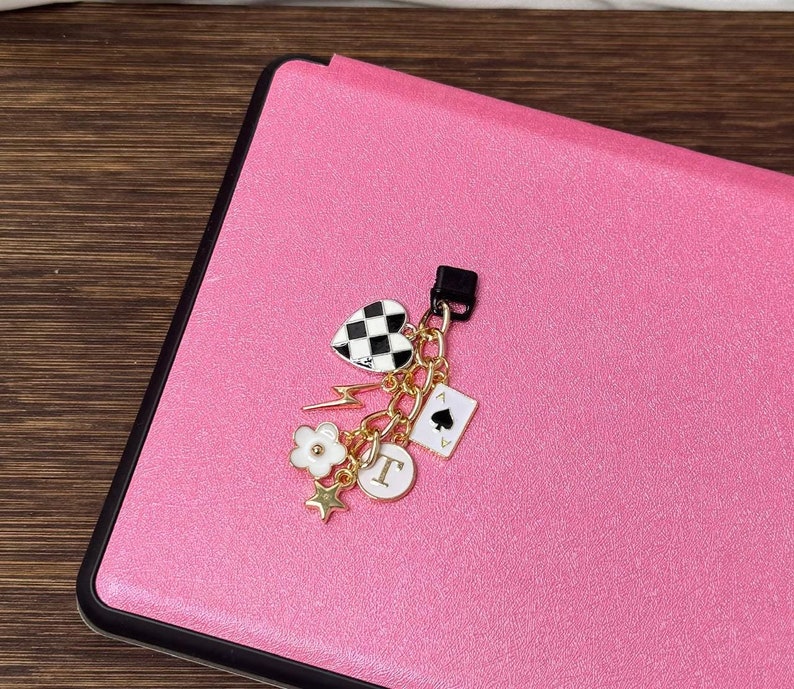 Op de afbeelding: Een roze leren notitieboekje met een gouden bedel eraan. De bedel heeft een zwart-wit geruit hart, een gouden bliksem, een witte bloem, een gouden ster, een gouden letter 'T' en een speelkaart met een schoppen symbool.
