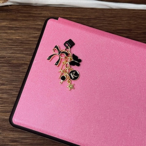 Op de afbeelding: Een roze leren notitieboekje met een gouden bedel eraan. De bedel heeft een zwarte strik, een gouden ster en een gouden letter 'K'.