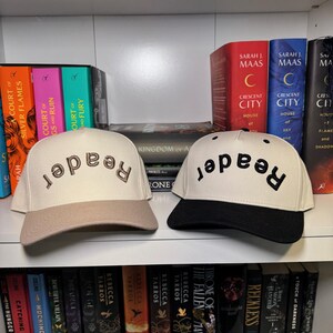 May include: Two baseball caps with the word "reader" embroidered on the front. One cap is beige with a brown brim, and the other is white with a black brim. The caps are displayed on a shelf with a collection of books.