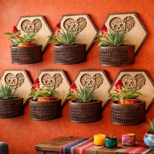 May include: Wall-mounted wooden planters with a Day of the Dead theme. Each planter features a laser-cut skull and heart design, holding small plants and flowers. The planters are arranged on an orange wall.