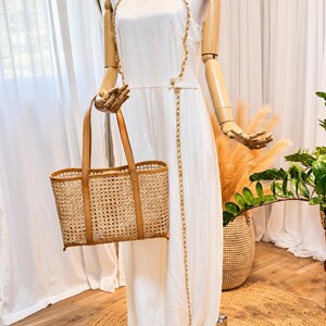 May include: A sleeveless white jumpsuit featuring a high neckline and gold chain straps that extend down the front. The garment is paired with a rectangular woven rattan tote bag with tan leather handles and trim, and light-coloured thong sandals.