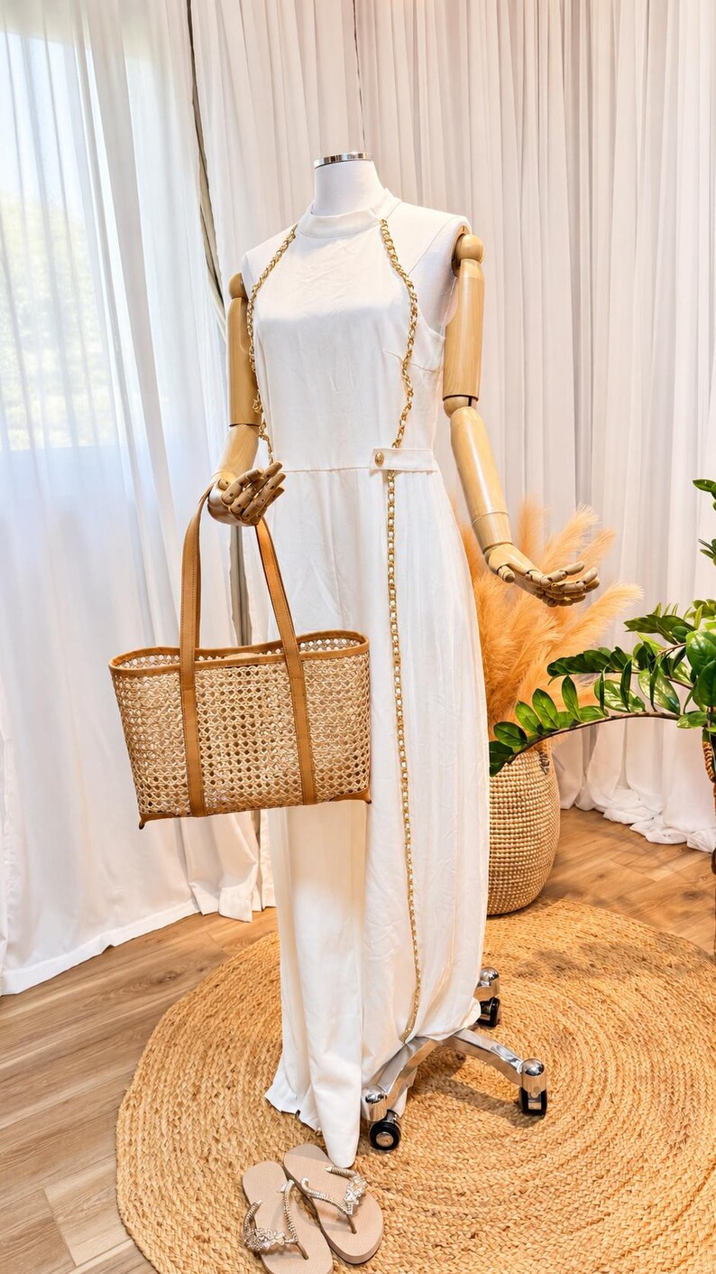 May include: A sleeveless white jumpsuit featuring a high neckline and gold chain straps that extend down the front. The garment is paired with a rectangular woven rattan tote bag with tan leather handles and trim, and light-coloured thong sandals.