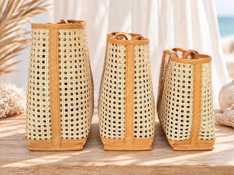 May include: Three tote bags of varying heights arranged in a row, each constructed with a light beige woven cane mesh body and tan leather trim along the vertical edges, top opening, and base, featuring two short handles on each bag.