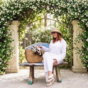 A woman in a white outfit and hat sits on a bench in a garden, holding a woven bag.